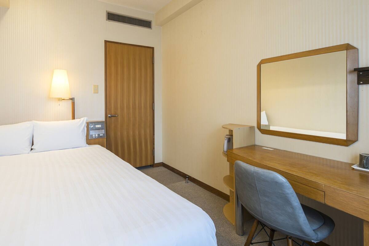 Habitación de hotel con cama, escritorio, silla, lámpara, espejo y puerta.