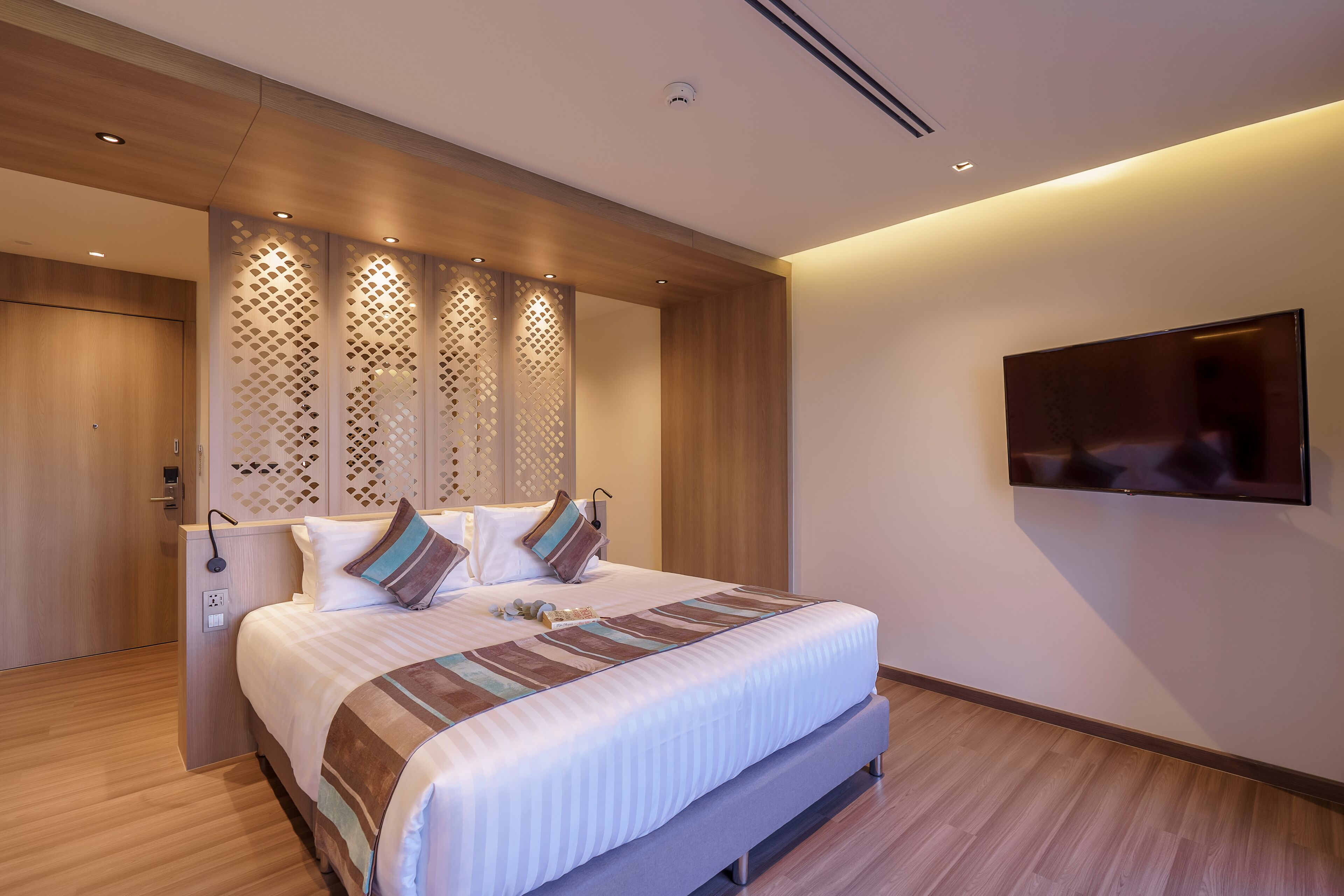 A modern hotel room with a large bed, wooden paneling, a TV mounted on the wall, and a power outlet on the side of the bed.