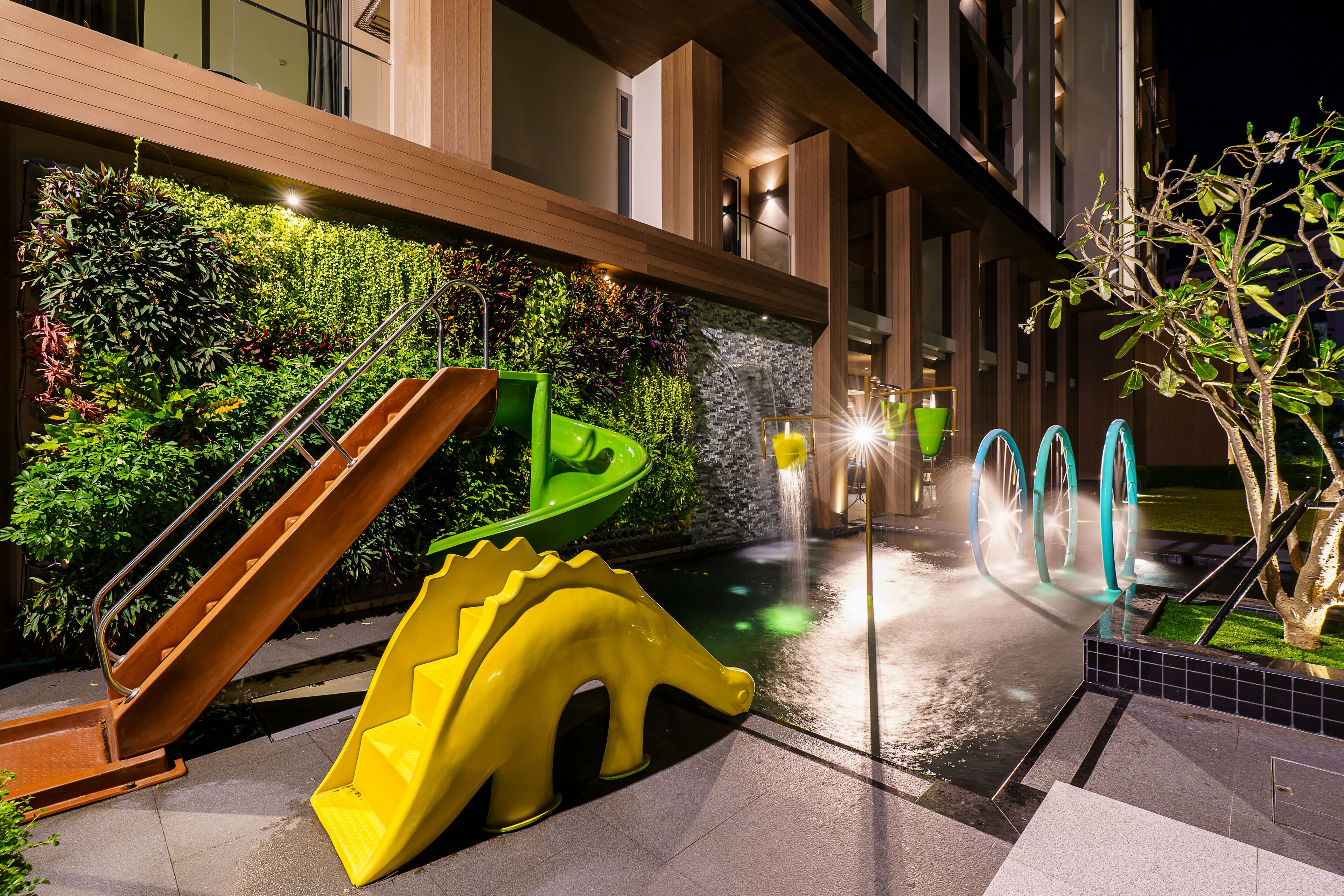 A modern building with a green slide, a yellow dinosaur-shaped play structure, and a water feature.