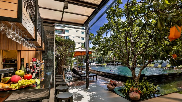 Uma área de bar com exposição de frutas, um espaço de lounge à beira da piscina e vista para um edifício ao fundo.