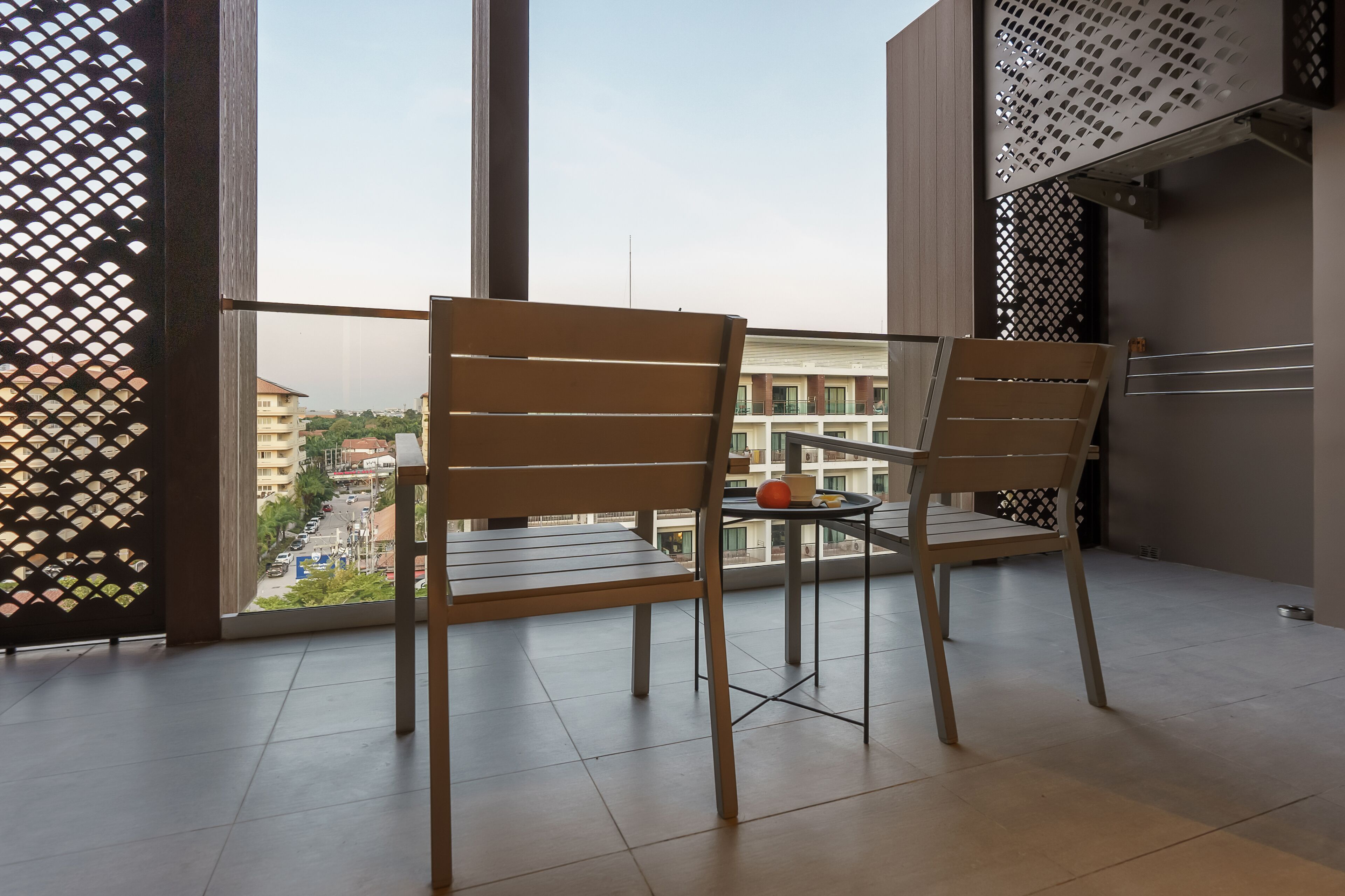 A balcony with two wooden chairs and a small table, a view of a building across the street, and a metal railing.