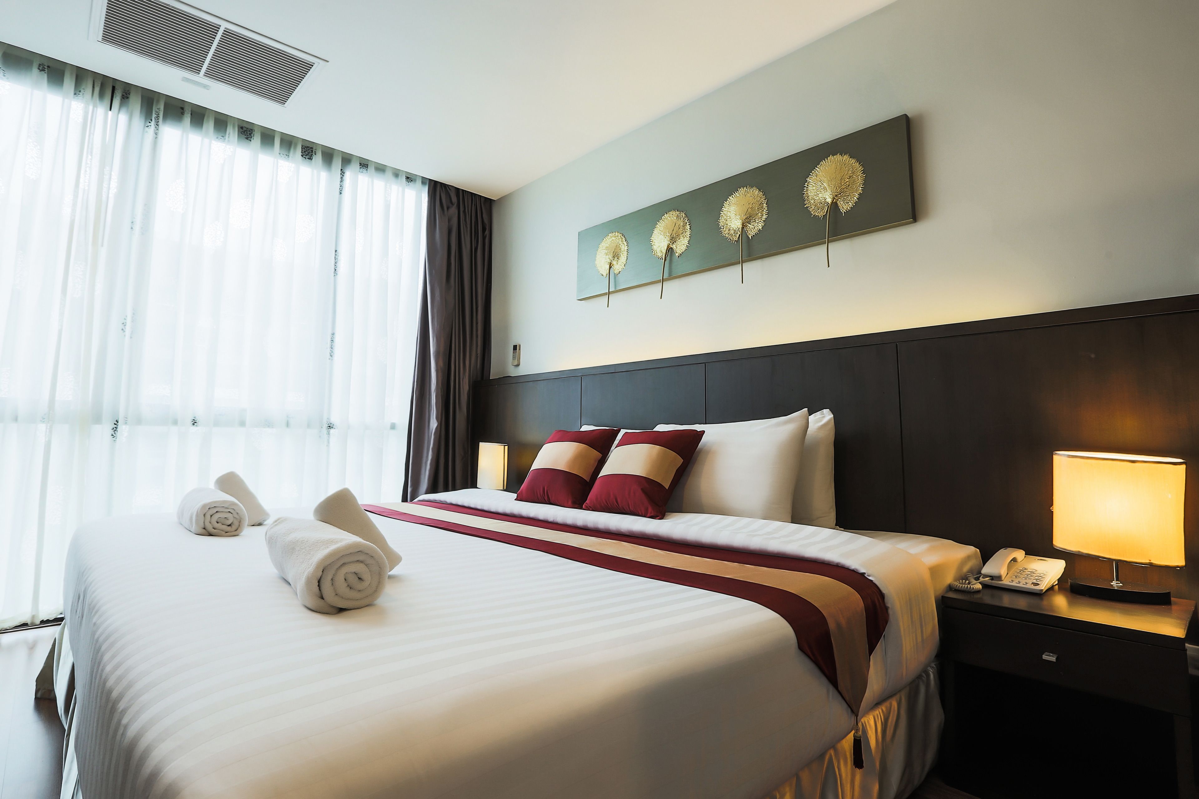 A neatly made bed with white linens and red accents, a bedside table with a lamp, and a wall-mounted artwork of three flowers.