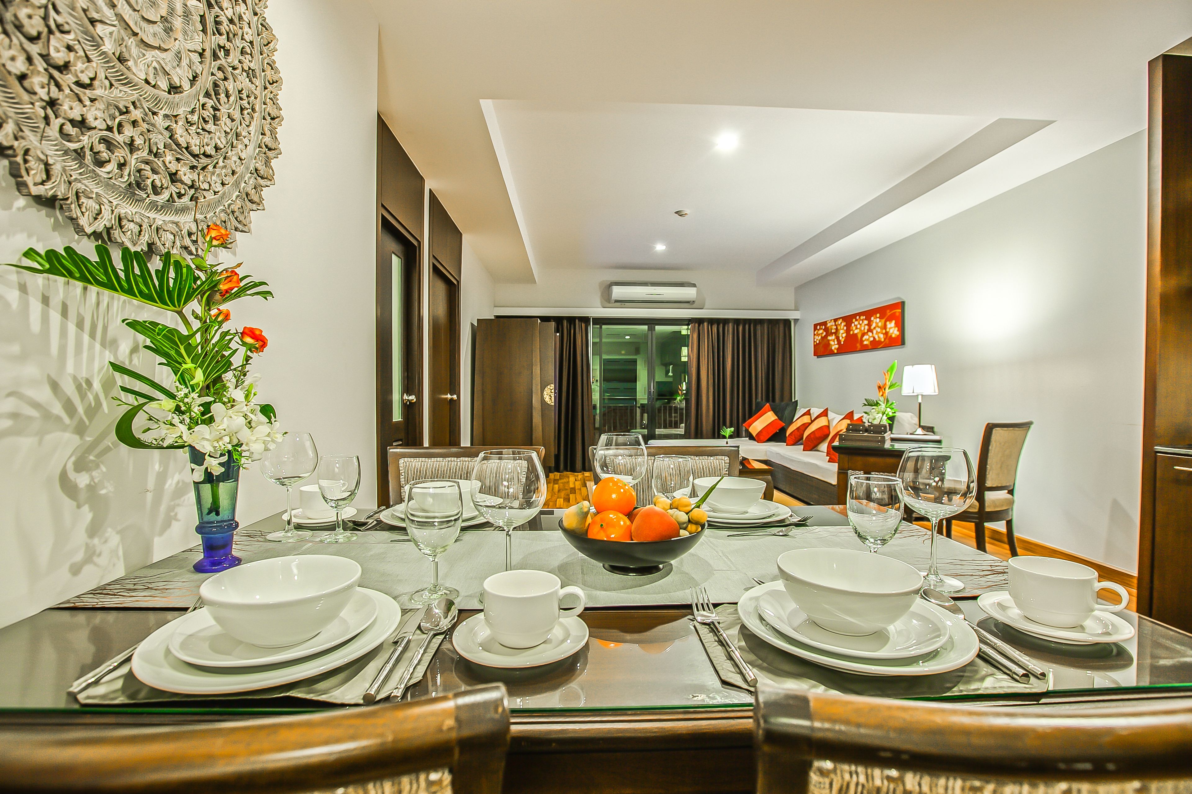 A dining area with a glass table set for a meal, a decorative wall, and a view into the living area.