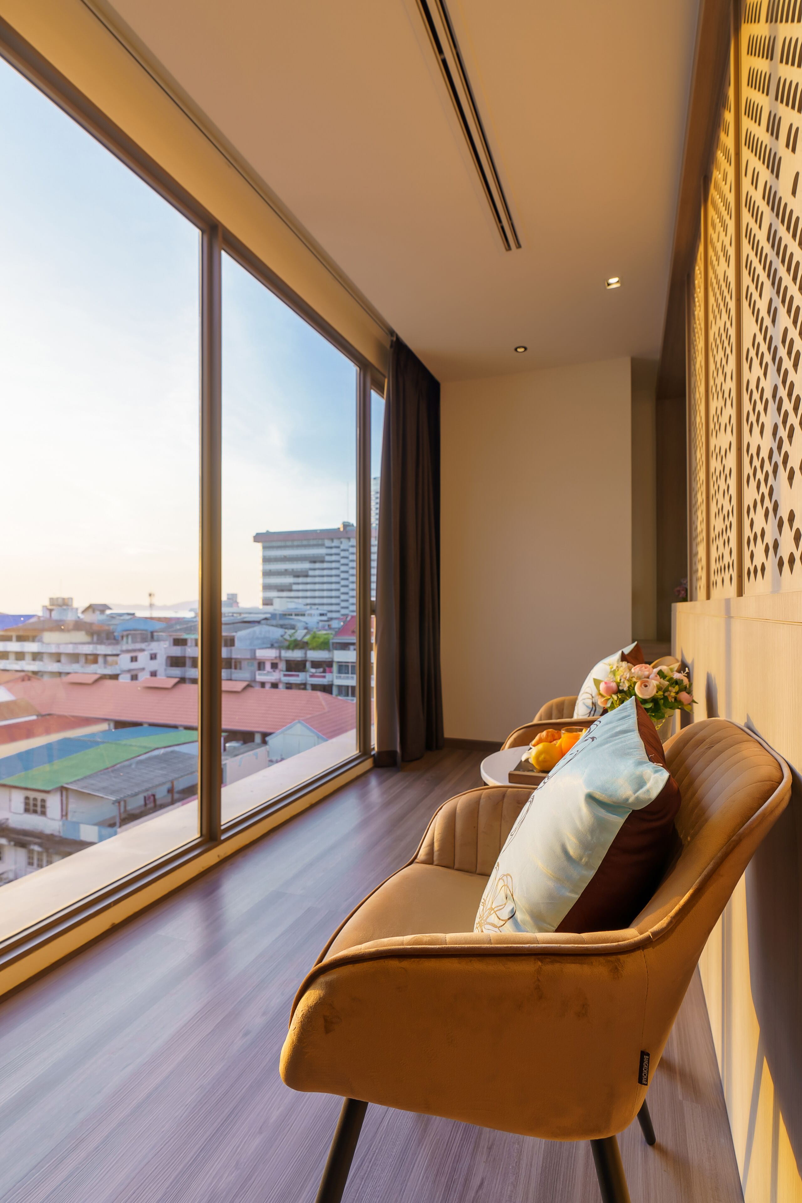 A modern hotel room with a large window offering a city view, a comfortable armchair with a decorative pillow, and a small table with a vase of flowers.