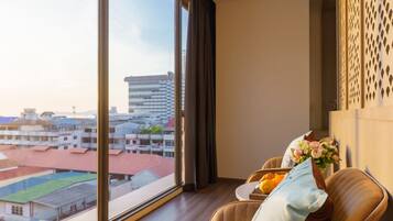 A modern hotel room with a large window offering a city view, a comfortable armchair with a decorative pillow, and a small table with a vase of flowers.