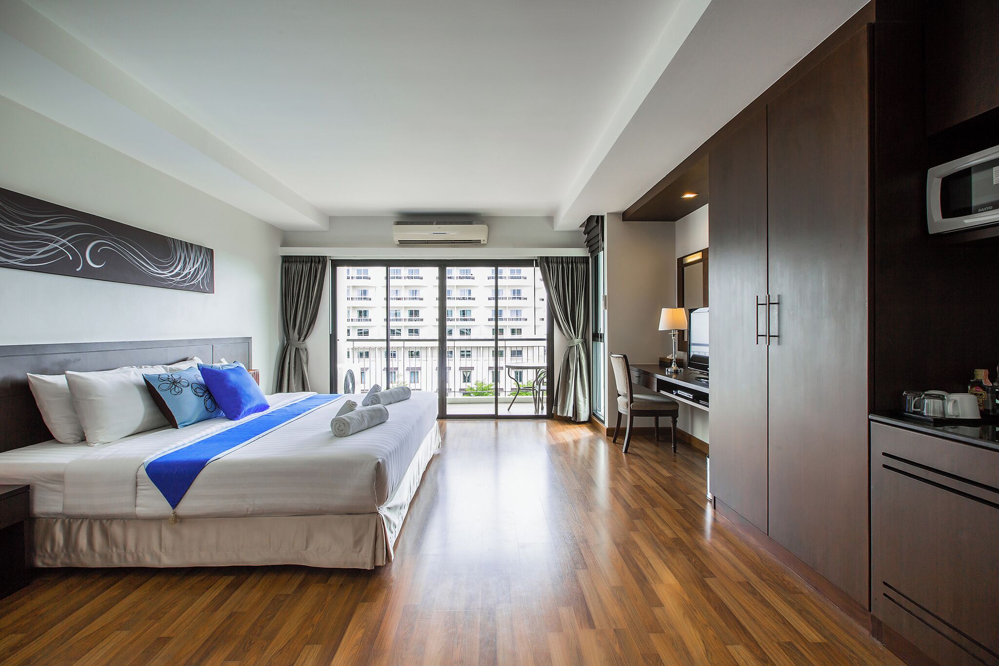 A hotel room with a bed, a desk, a microwave, and a view of a building through large windows.