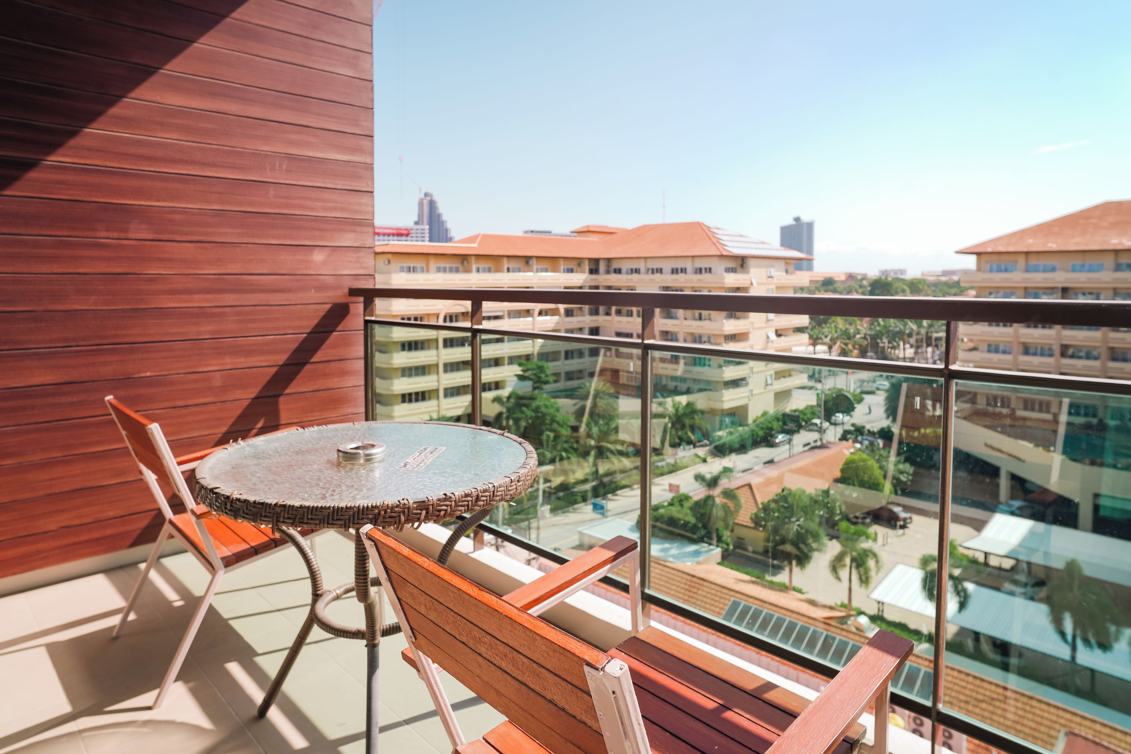 Un balcon avec une table ronde en verre et deux chaises en bois, donnant sur un paysage urbain avec des bâtiments et un ciel dégagé.