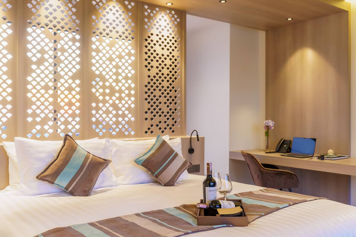 A hotel room with a bed, a desk, and a laptop. There is a bottle of wine and a glass on the bed.