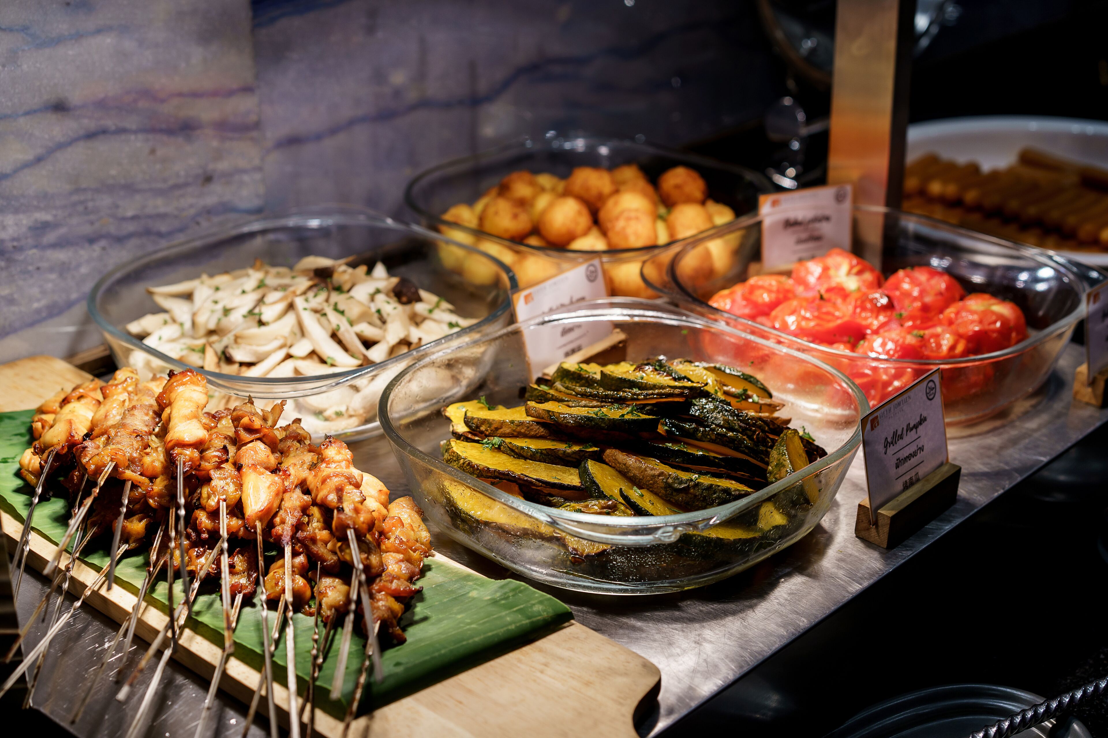 A buffet with various food options including skewered meat, mushrooms, roasted potatoes, and sliced vegetables.