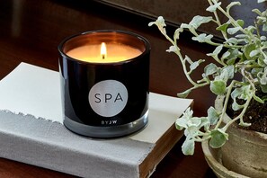 A black candle with "SPA" and "BYJW" on it, placed on a white cloth next to a book and a potted plant. - The Headquarters Inn (Nairobi)