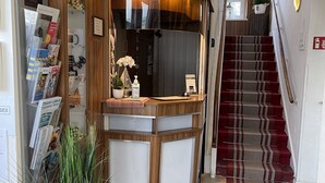 Un hall d’accueil d’hôtel avec un comptoir de réception en bois, un panneau d’accueil dans plusieurs langues, un escalier et un paillasson rouge orné des initiales de l’hôtel.