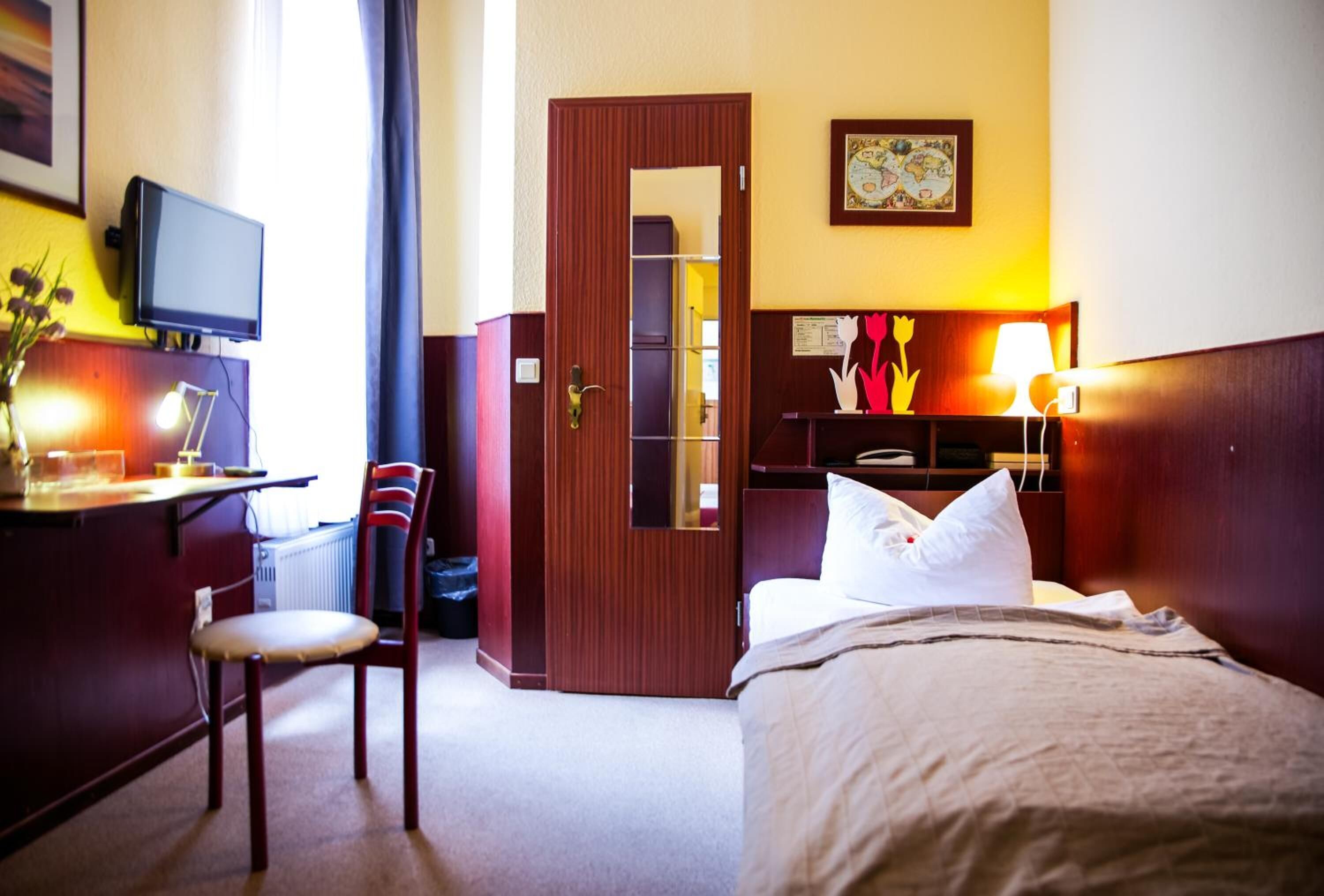 Una habitación de hotel con una cama, un escritorio con televisor, una silla, un armario y una mesita de noche con lámpara.