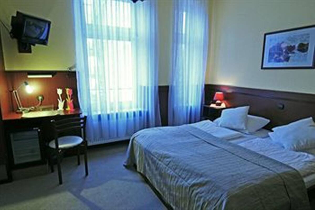 A hotel room with a bed, a desk, a chair, a television mounted on the wall, and a window with curtains.