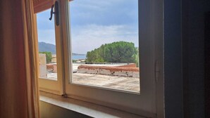 A view through a window with a metal railing, a concrete floor, and a distant view of trees and a body of water. - King's Hotel (Golfo Aranci)