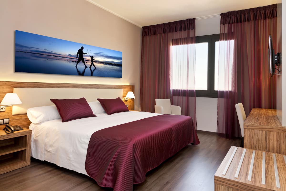 A hotel room with a large bed, two bedside tables, a desk, and a chair. There is a painting on the wall above the bed.