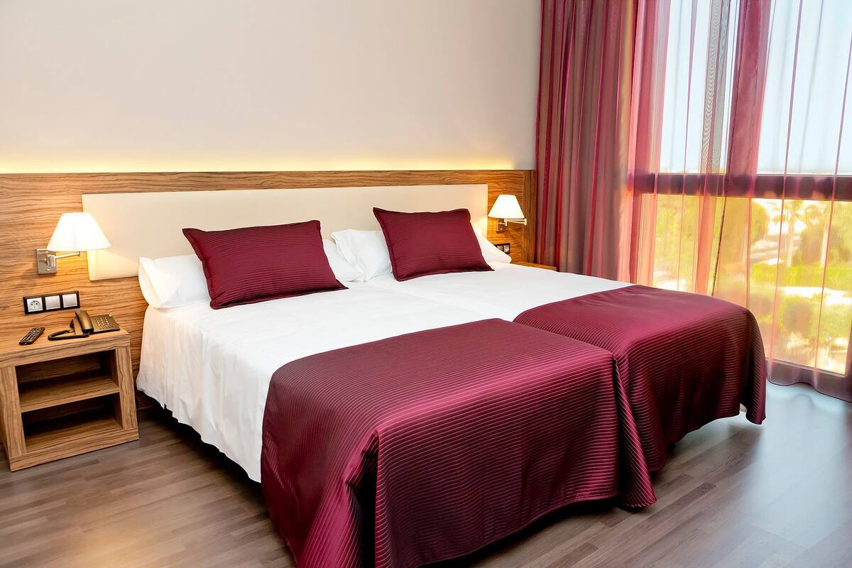 A double bed with two red pillows, a wooden nightstand with a lamp, and a view of greenery through sheer curtains.