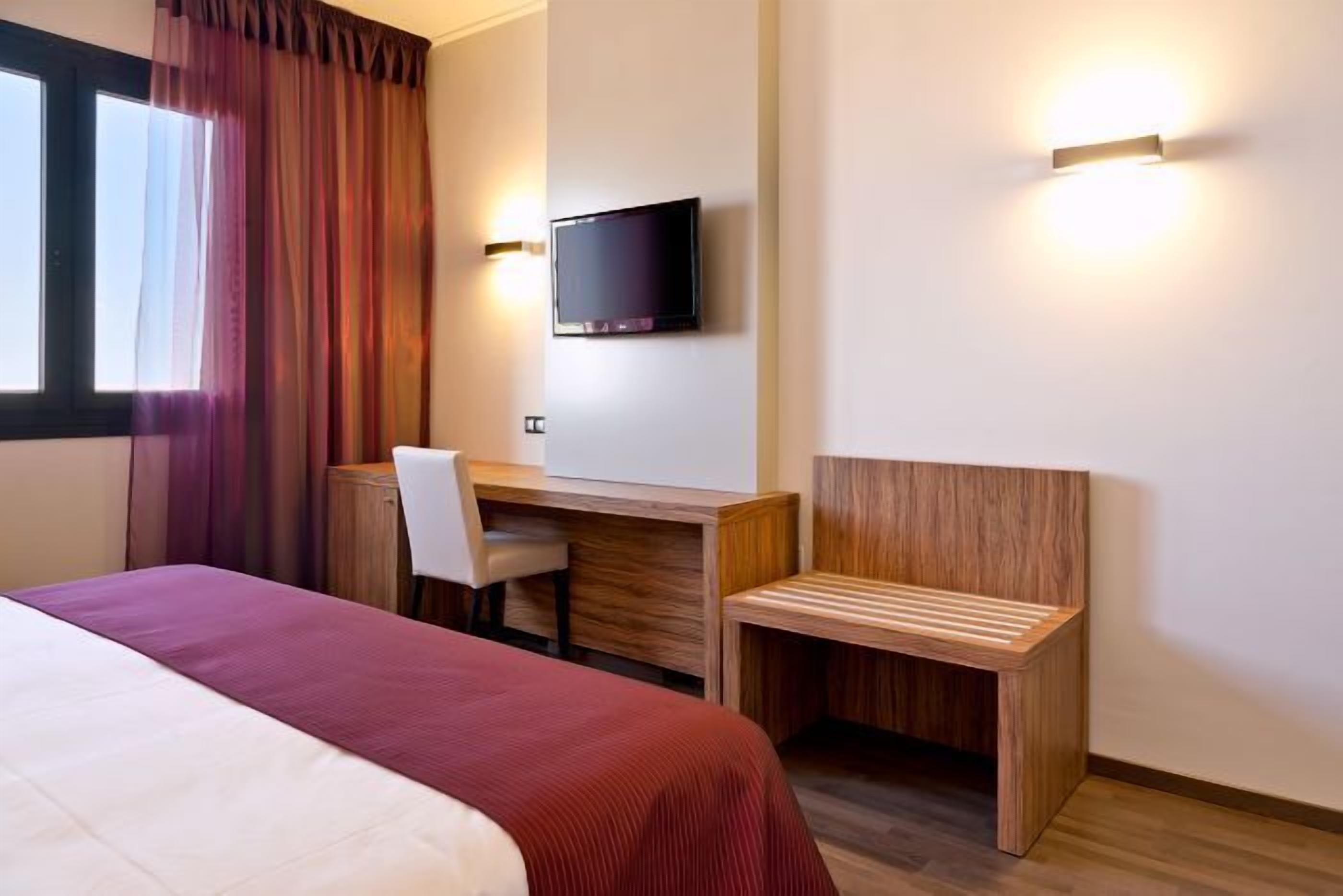 A hotel room with a bed, a desk, a chair, a TV, and a bench.