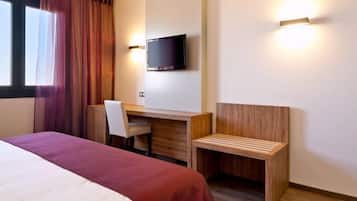 A hotel room with a bed, a desk, a chair, a TV, and a bench.