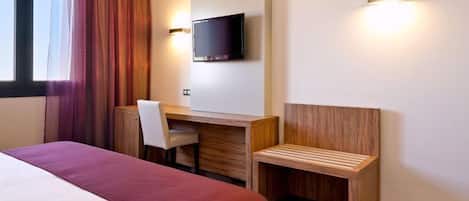 A hotel room with a bed, a desk, a chair, a TV, and a bench.
