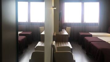 A hotel room with a large bed, a desk, and two chairs. There are two windows with curtains, and the room has a wooden floor.