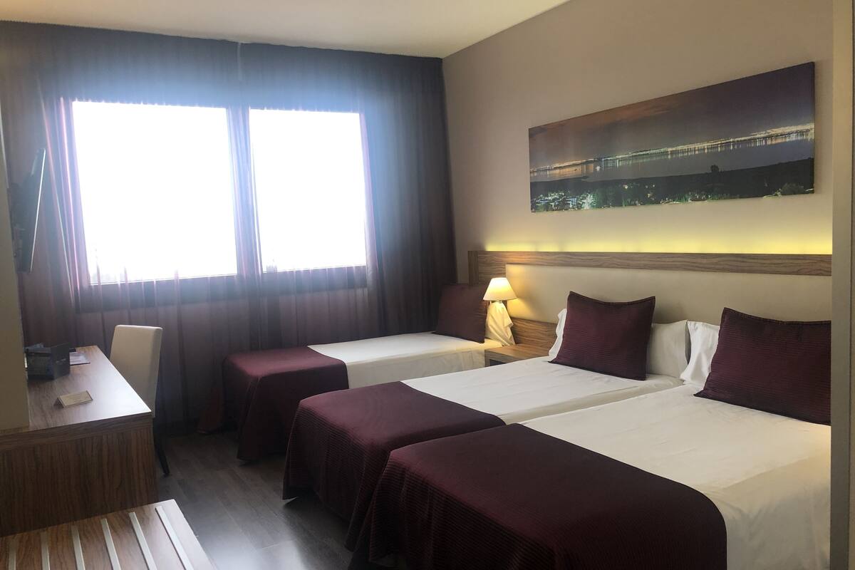 A hotel room with two beds, a desk, and a large window.