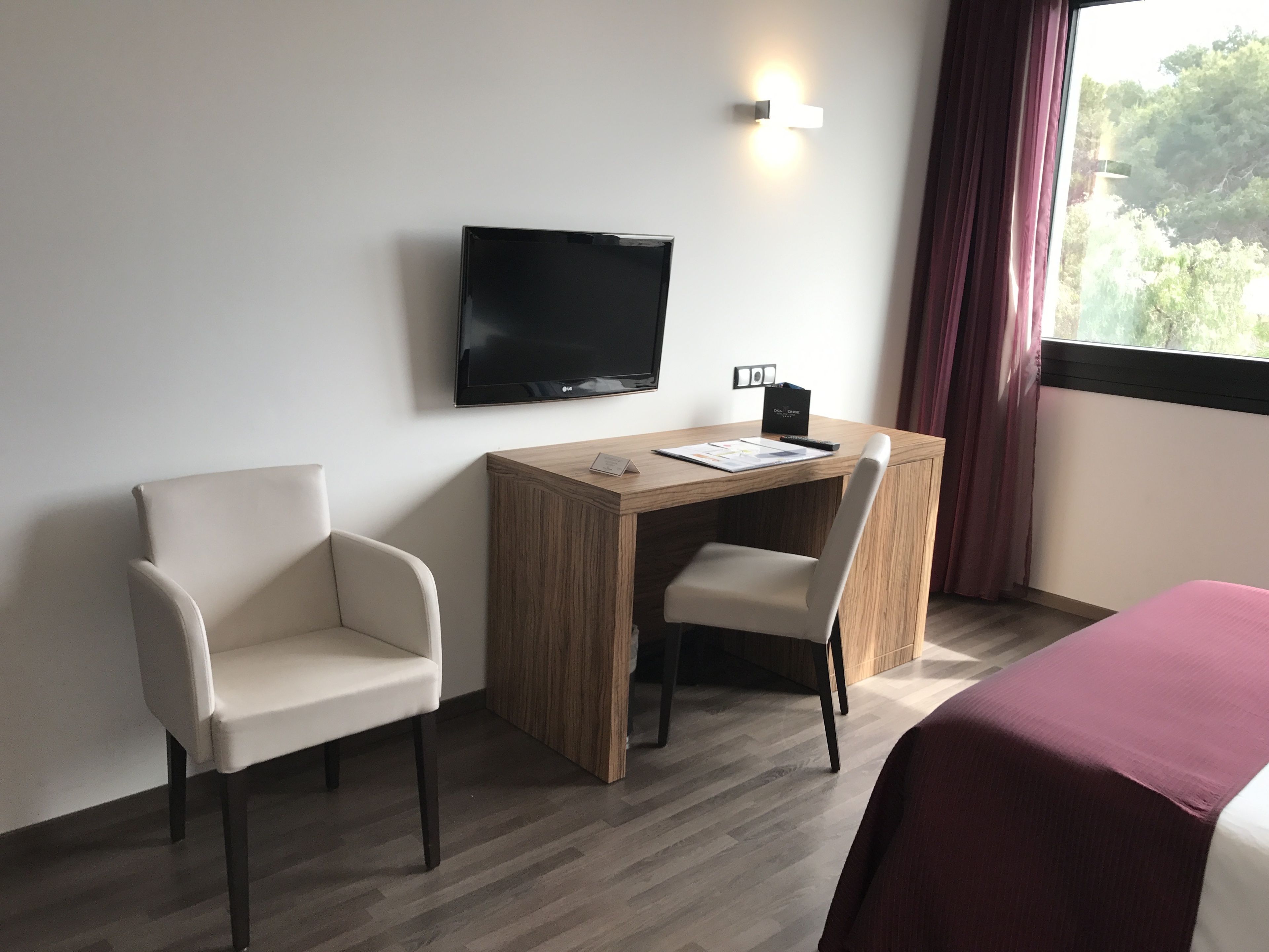 Een hotelkamer met een houten bureau, twee stoelen, een televisie en een bed met een rode spreidekker.