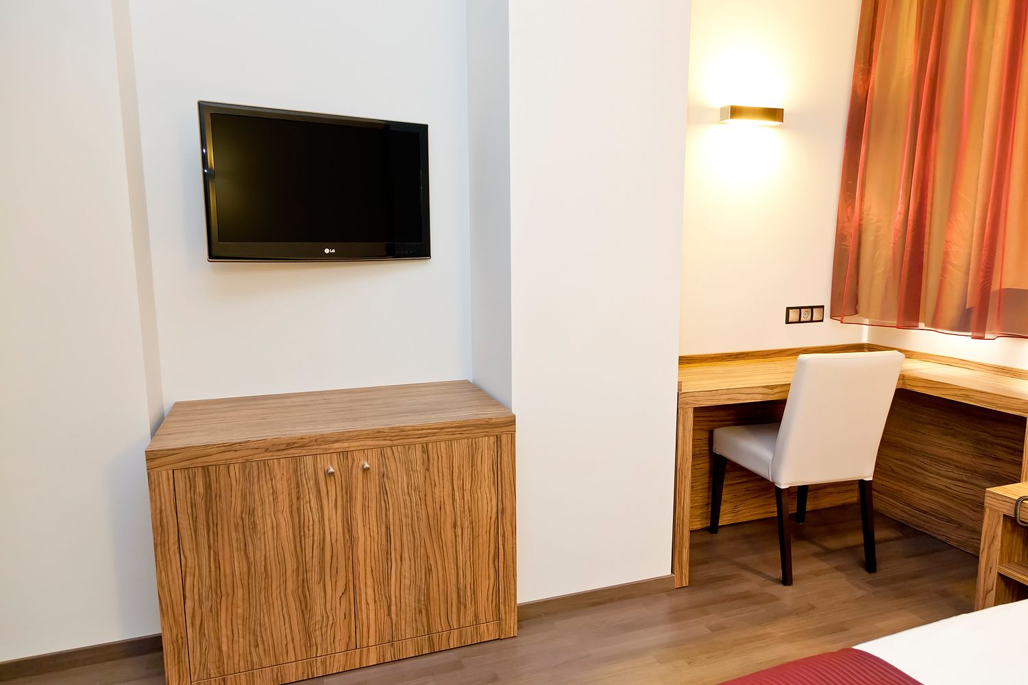A hotel room with a wooden desk, a chair, a flat-screen TV mounted on the wall, and a bedside table.