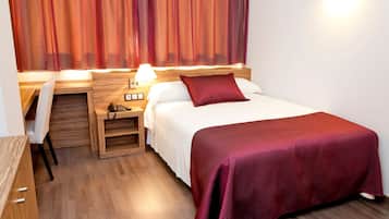 A hotel room with a bed, a desk, and a chair. There is a lamp on the desk and a phone on the nightstand.