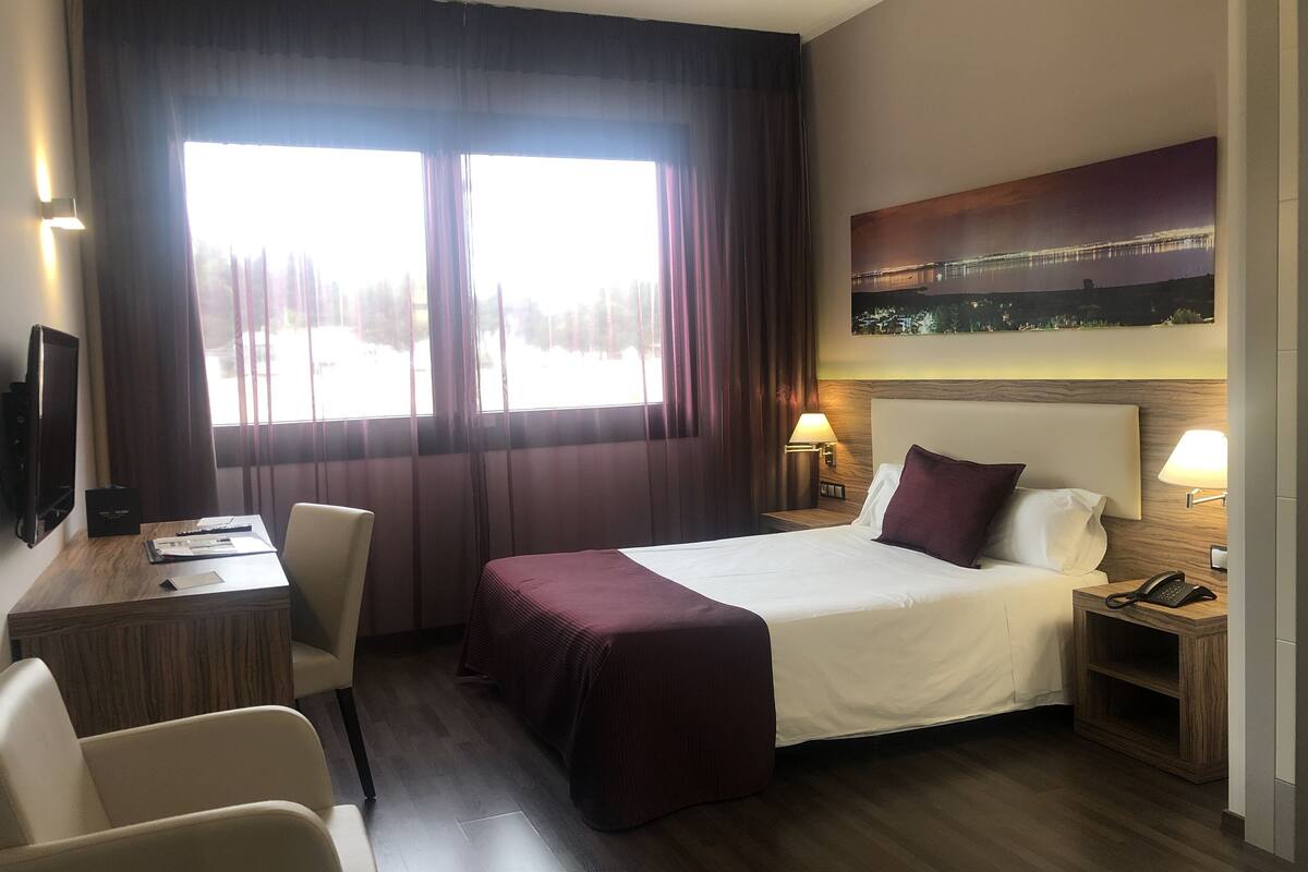 A hotel room with a large bed, a desk, a chair, a nightstand with a phone, and a painting on the wall.
