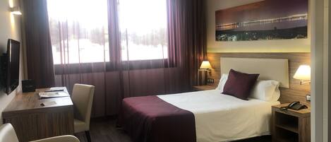 A hotel room with a large bed, a desk, a chair, a nightstand with a phone, and a painting on the wall.