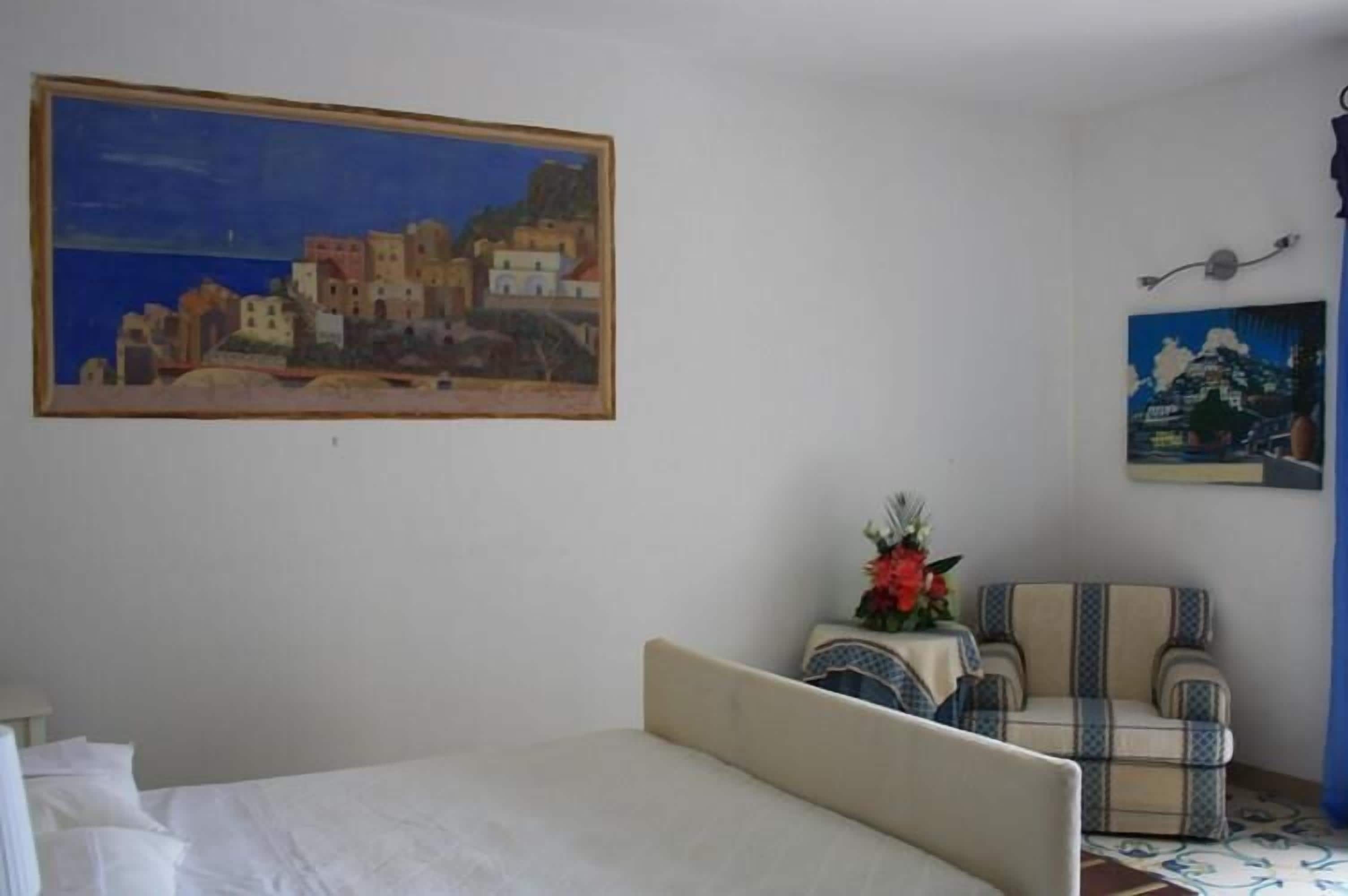 Un dormitorio con una cama, una silla, una mesita con un florero y dos cuadros en la pared.