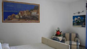 Un dormitorio con una cama, una silla, una mesita con un florero y dos cuadros en la pared.