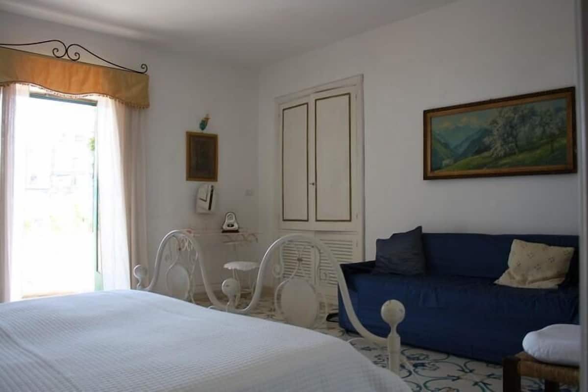 A bedroom with a bed, a blue sofa, a white chair, a small table, and a painting on the wall.