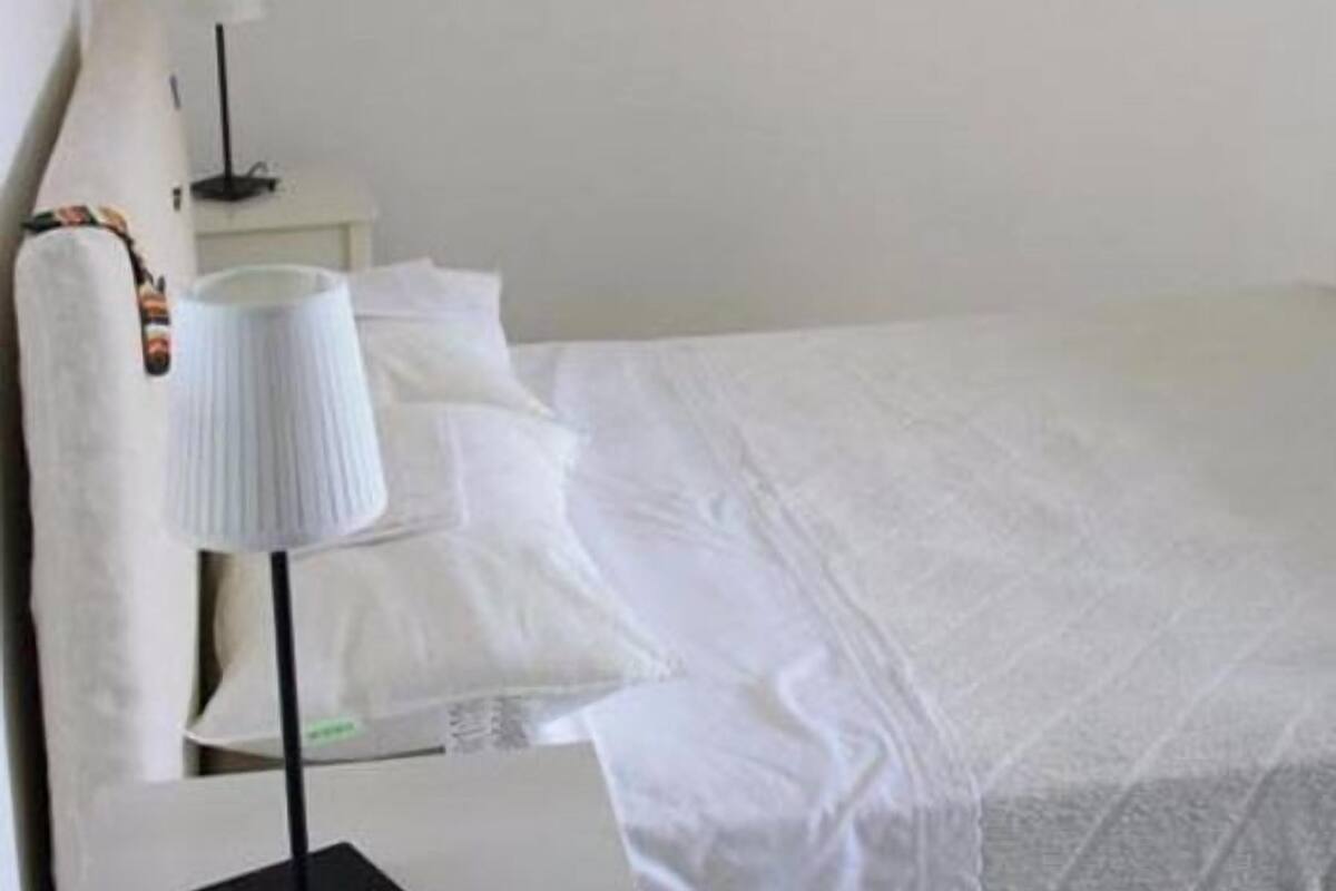 A hotel room with a bed, a bedside table with a lamp, a painting of a coastal town, and a wall-mounted lamp.