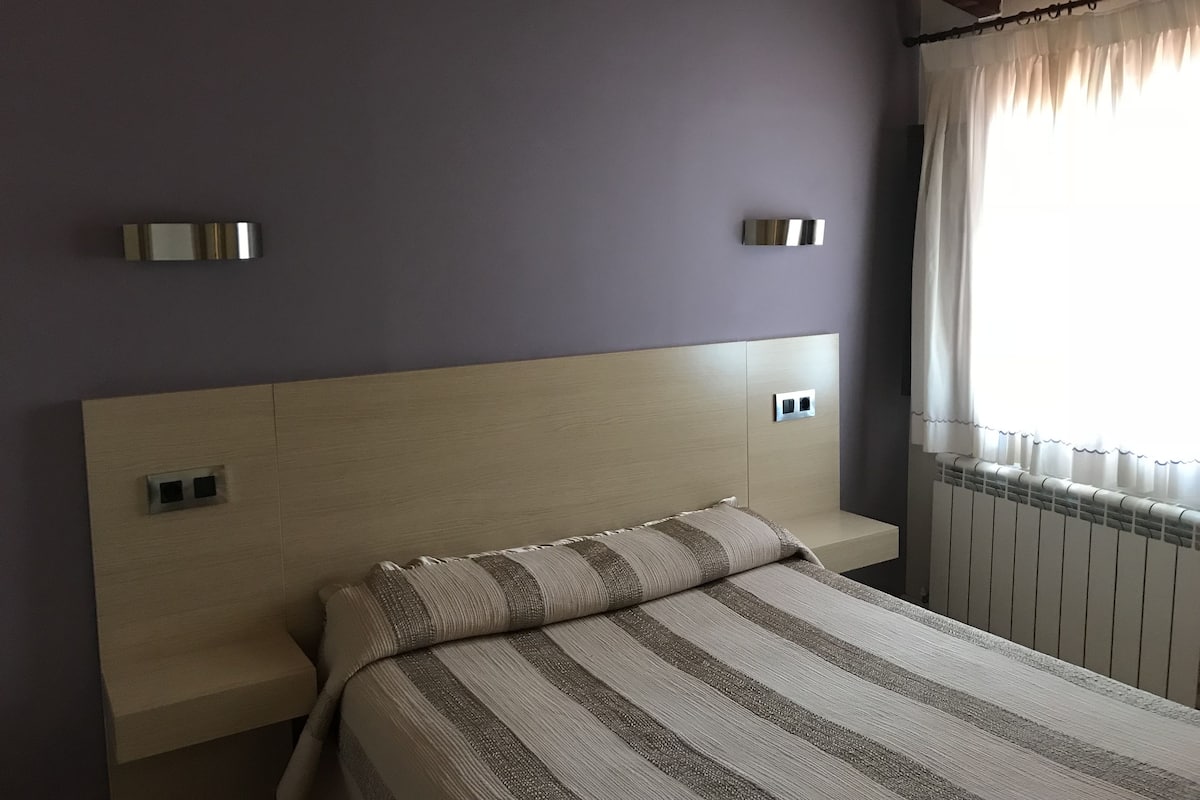 Un dormitorio con cabecera de madera, colcha a rayas y ventana con cortinas.