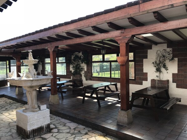 An outdoor patio with stone flooring, wooden tables and benches, a decorative fountain, and potted plants.