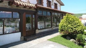 A two-story building with a tiled roof, large windows, and a small garden area with a bush. - Hotel Costa San Juan De La Canal (Santa Cruz de Bezana)