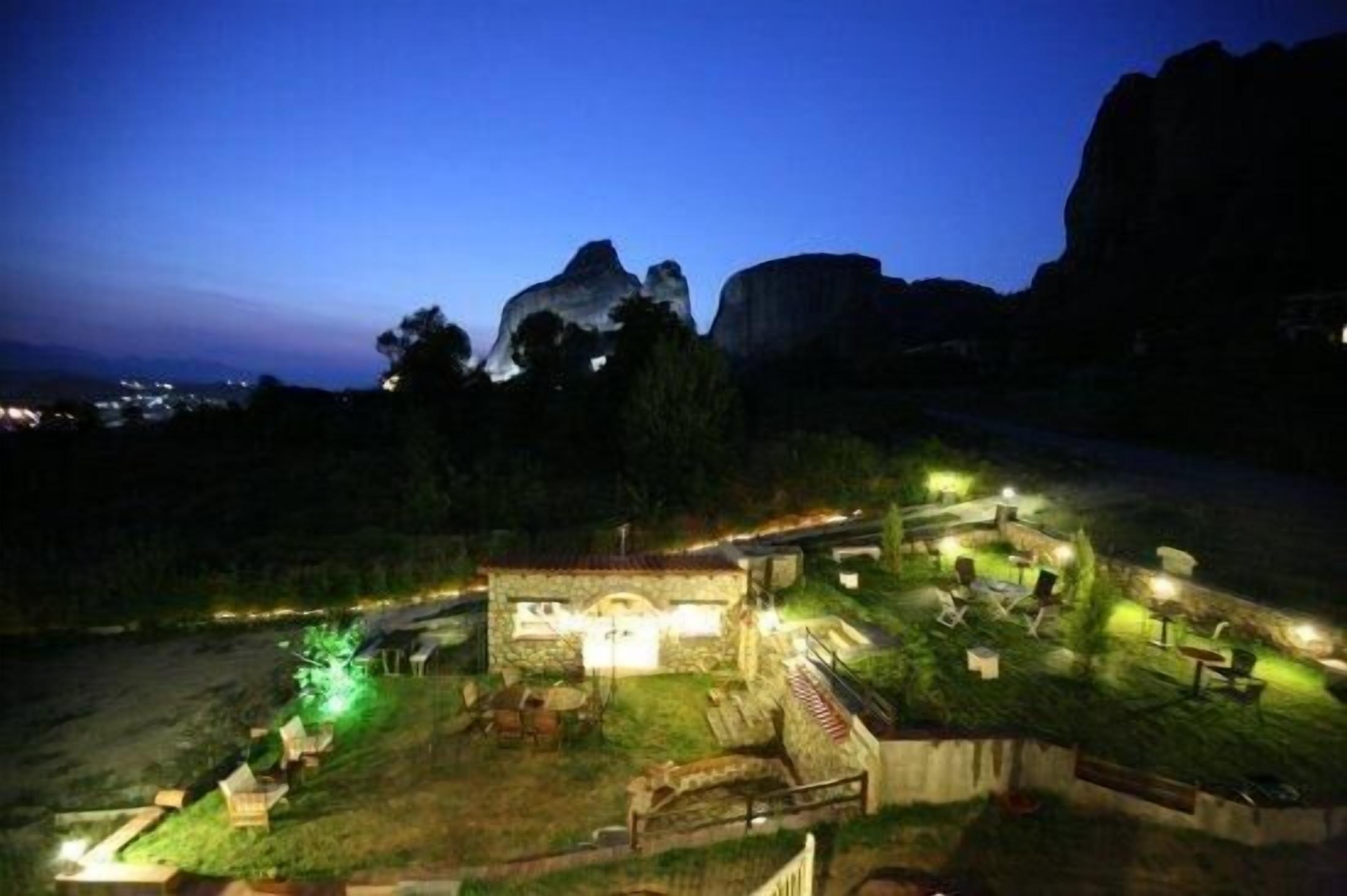 Vista nocturna de un refugio rústico con construcción de piedra, asientos al aire libre y vistas de una cadena montañosa.