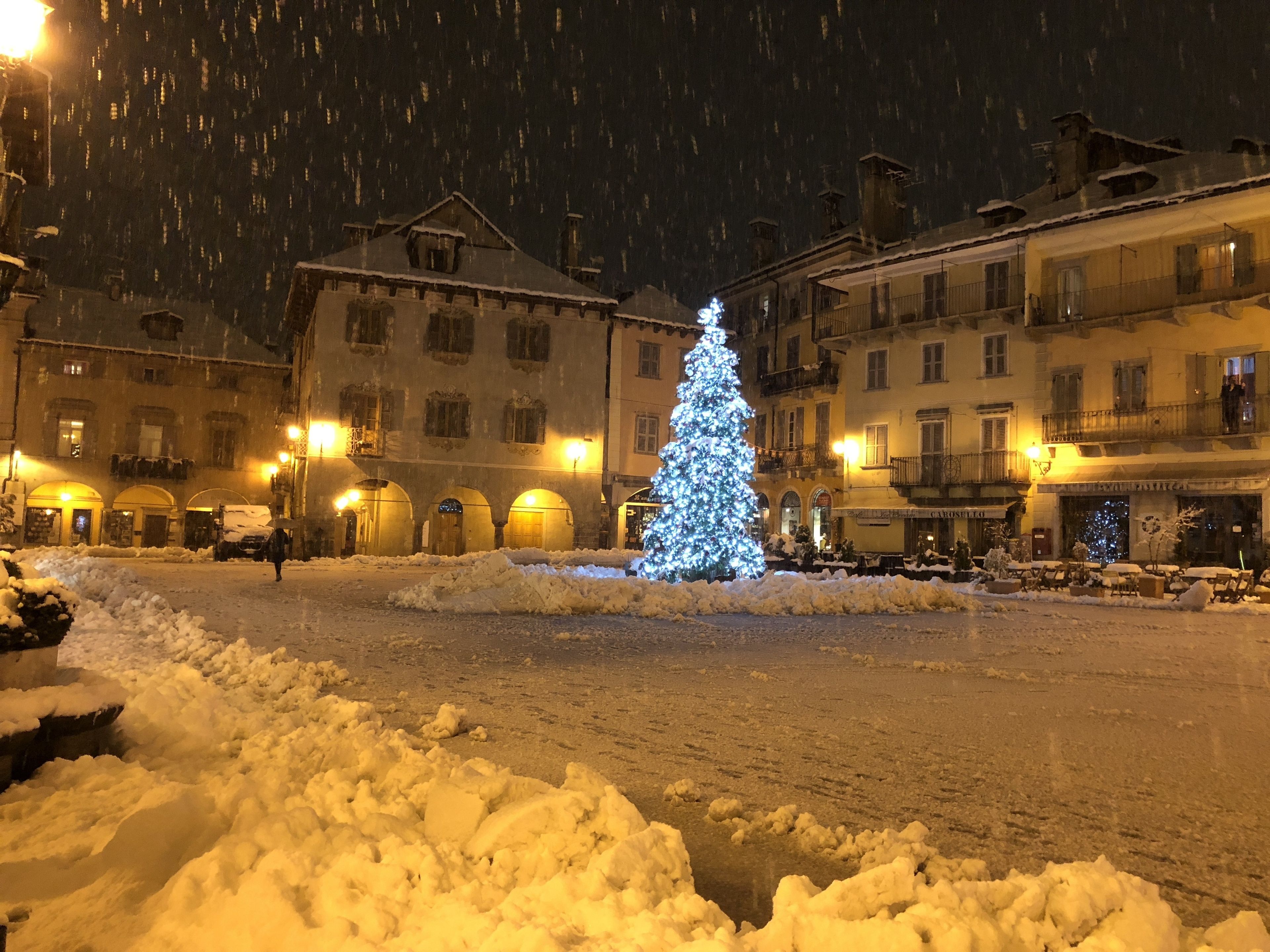 A snow-covered square with a lit Christmas tree in the center, surrounded by buildings with balconies and lit windows.