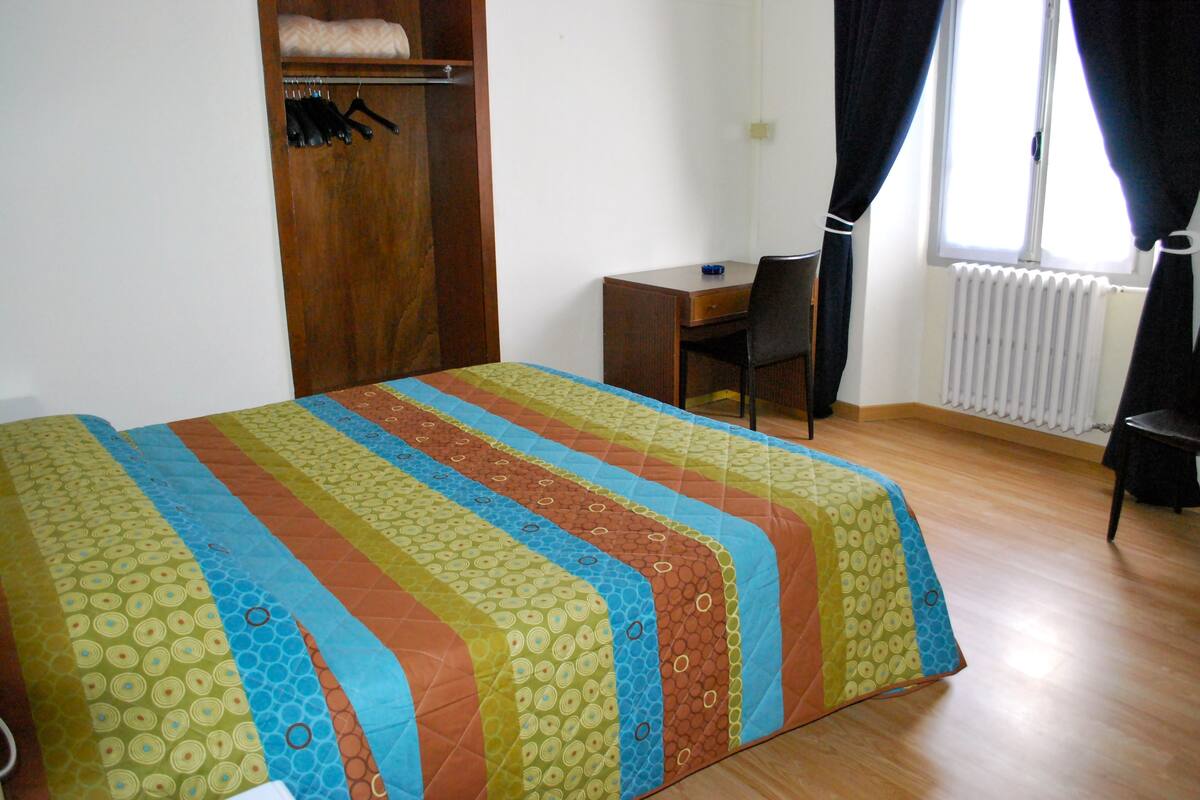 Une chambre à coucher avec un lit, un bureau et une chaise. On y trouve une armoire avec des cintres et un radiateur.