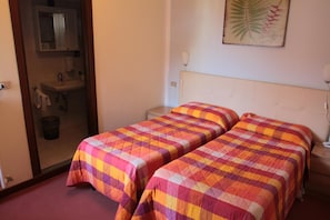 A room with two single beds, a small bedside table, a nightstand, a mirror, a sink, and a framed plant on the wall.