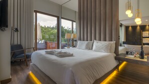 A modern hotel room with a large bed, a TV, a desk with a bottle and glasses, and a balcony with a view of trees.