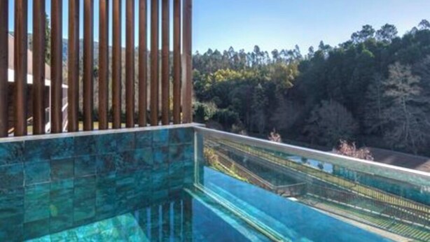 A modern outdoor infinity pool with a wooden pergola overhead, surrounded by a forested landscape.