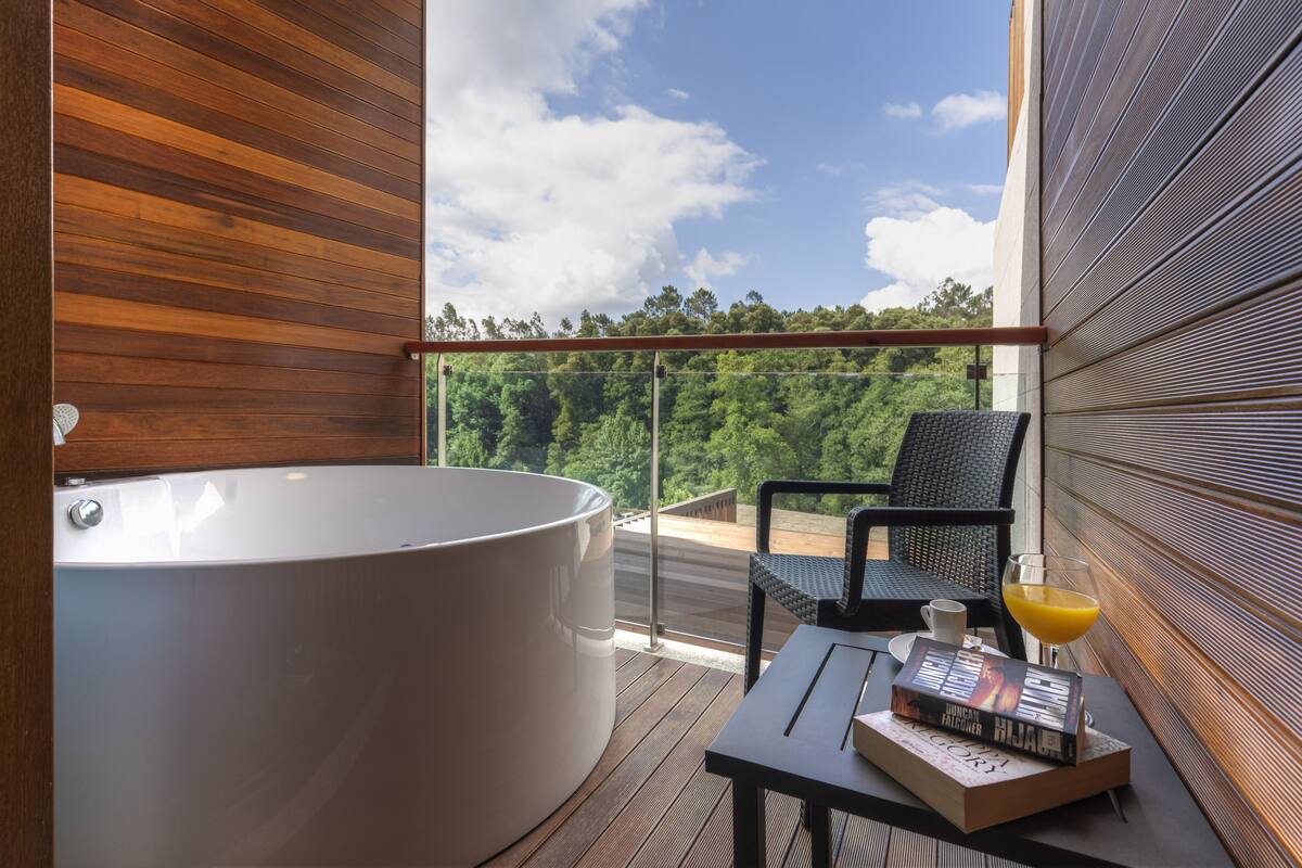 A modern bathroom with a large bathtub, a wooden floor, and a balcony with a view of the forest.