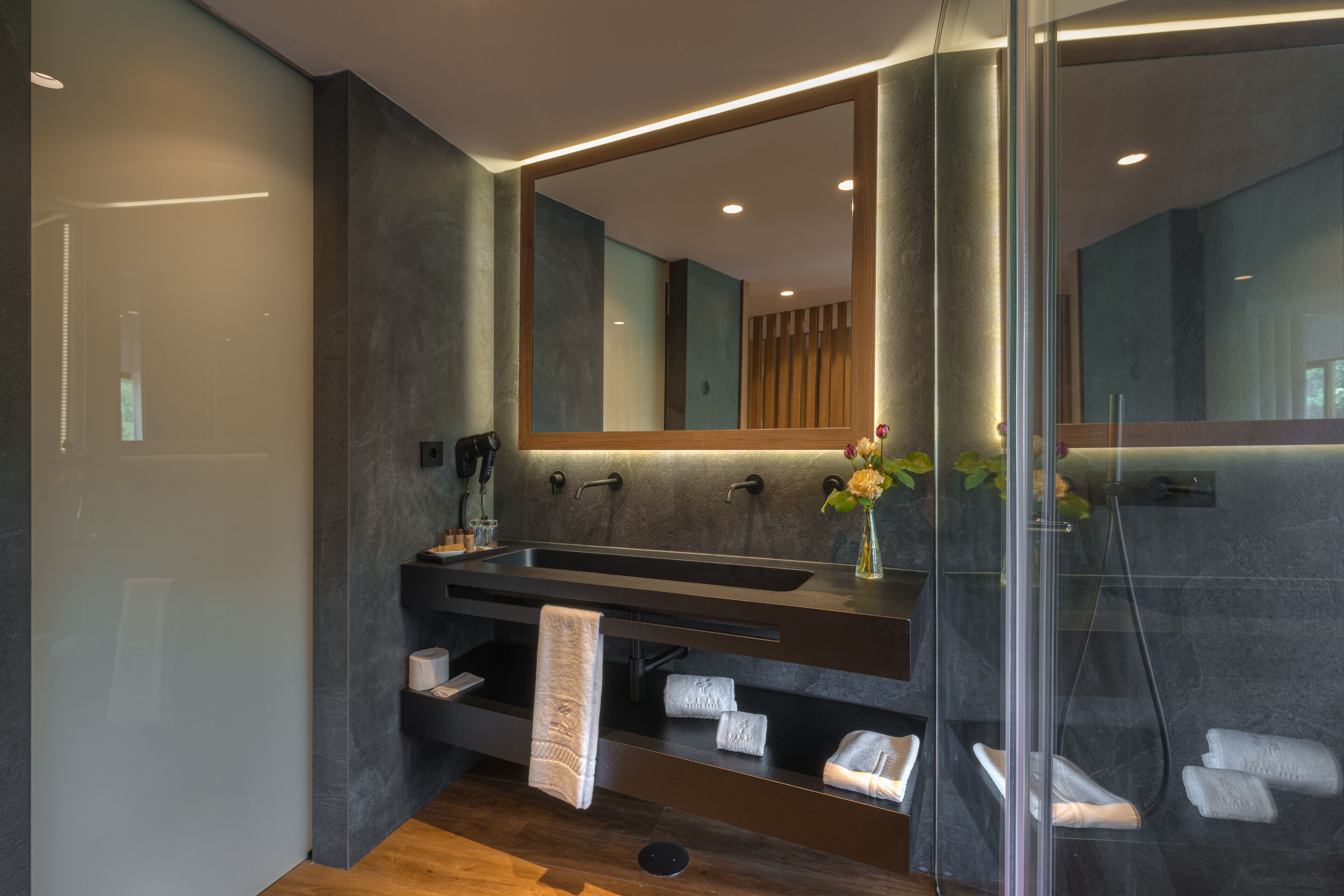 Une salle de bain moderne avec un grand miroir, des murs en pierre sombre, un meuble-lavabo en bois et une cabine de douche en verre.