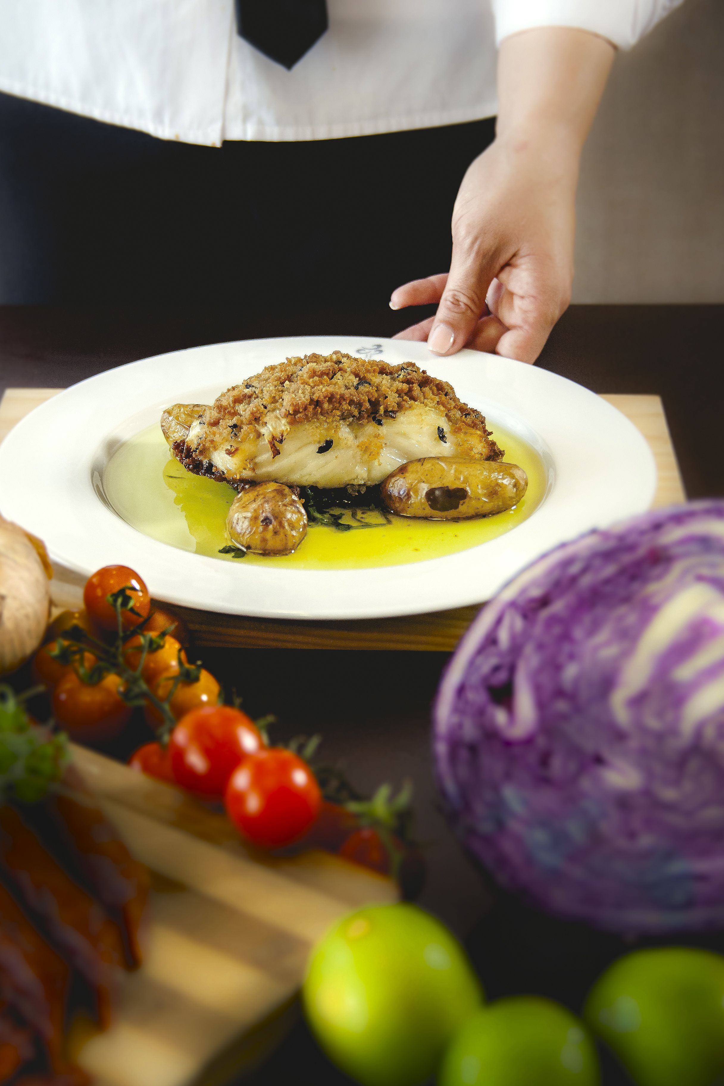 Ein Koch serviert ein Gericht mit goldbrauner Kruste, umgeben von frischen Gemüsesorten wie Kirschtomaten und Rotkohl.