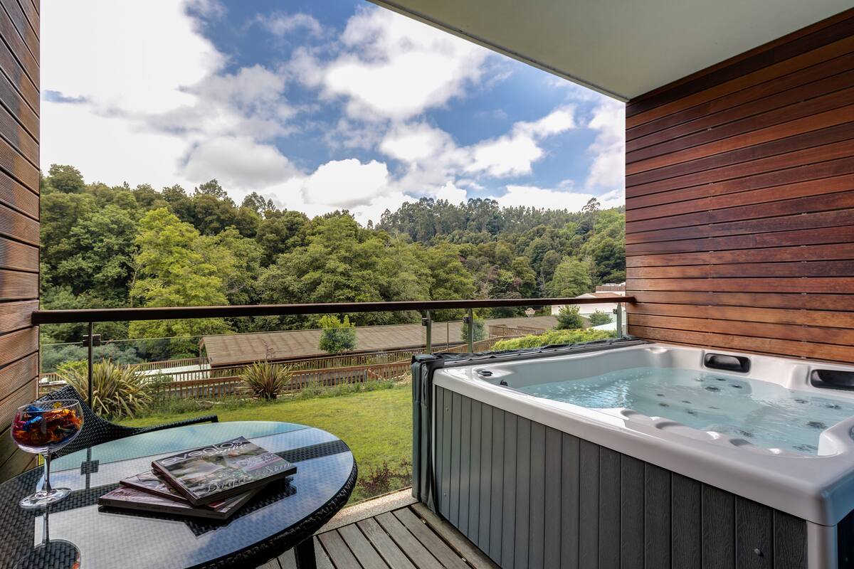 A balcony with a hot tub, a glass table with magazines, and a view of a forested area.