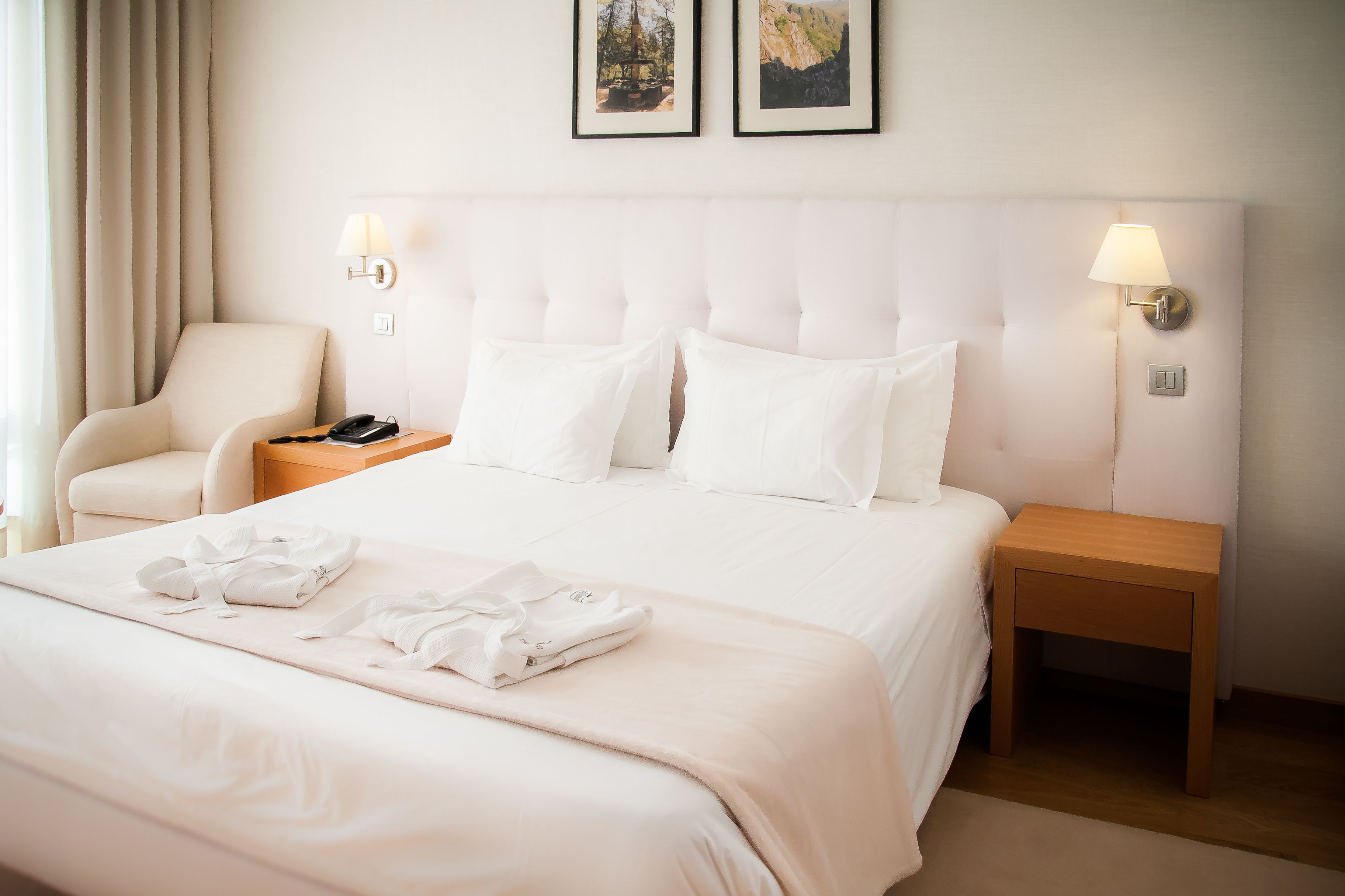 Una habitación de hotel con cama, una silla, una mesita de noche con lámpara y dos cuadros en la pared.