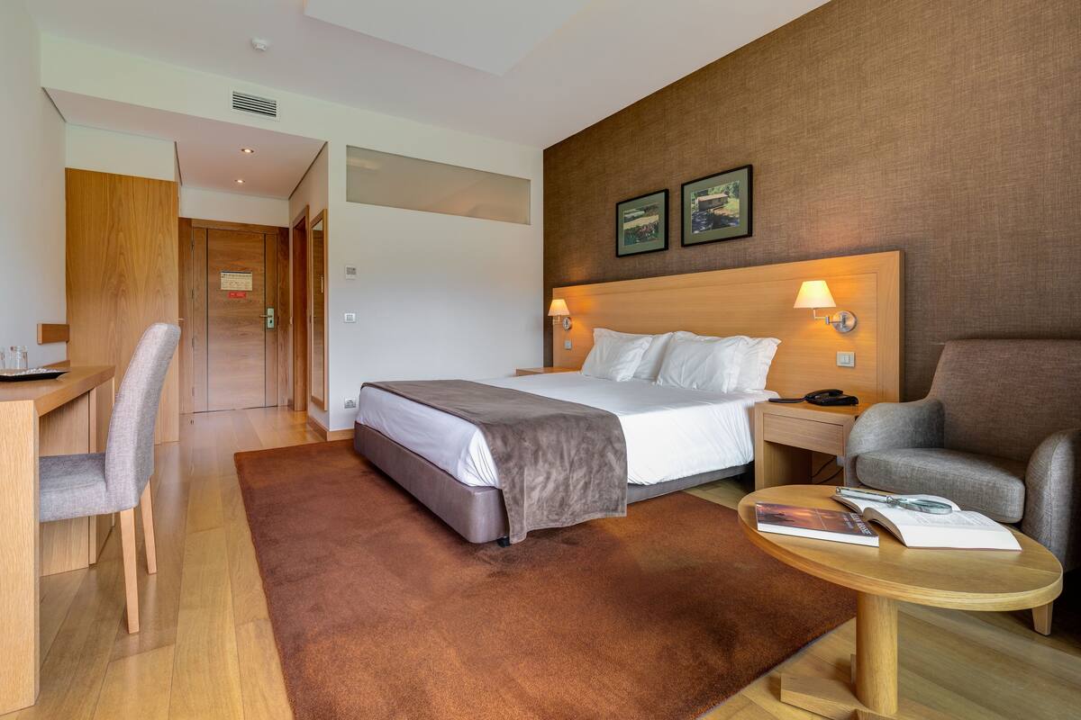 A hotel room with a bed, a desk, a chair, a small table, and a brown rug.