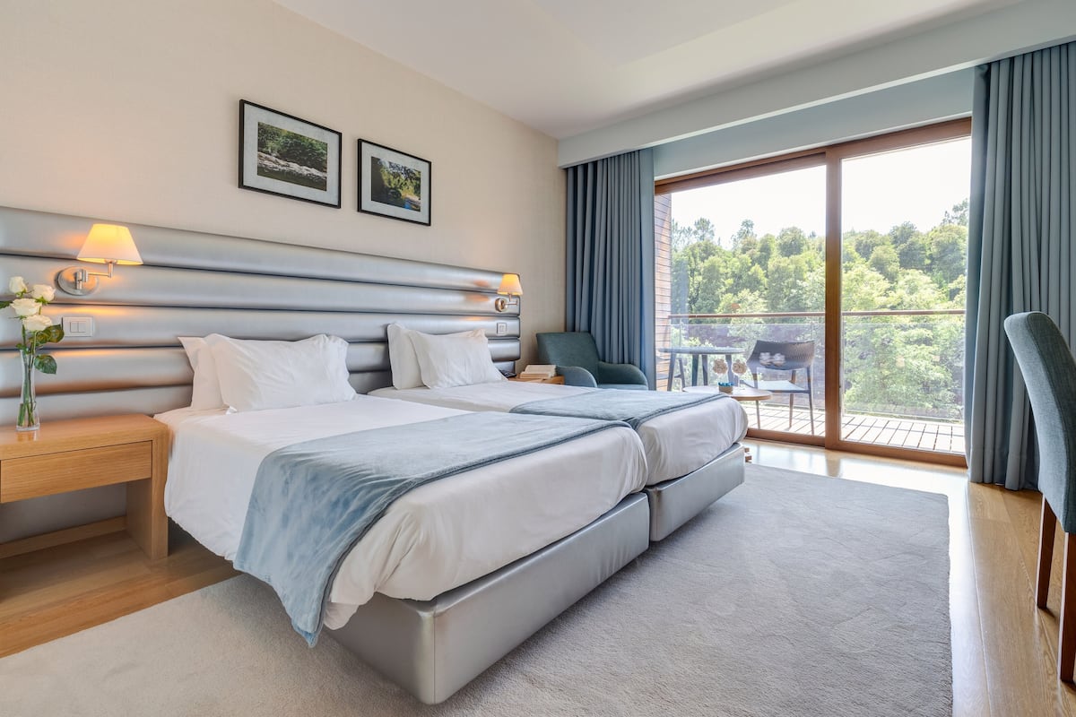 A modern hotel room with a large bed, two bedside tables, a nightstand, a chair, and a balcony with a view of trees.